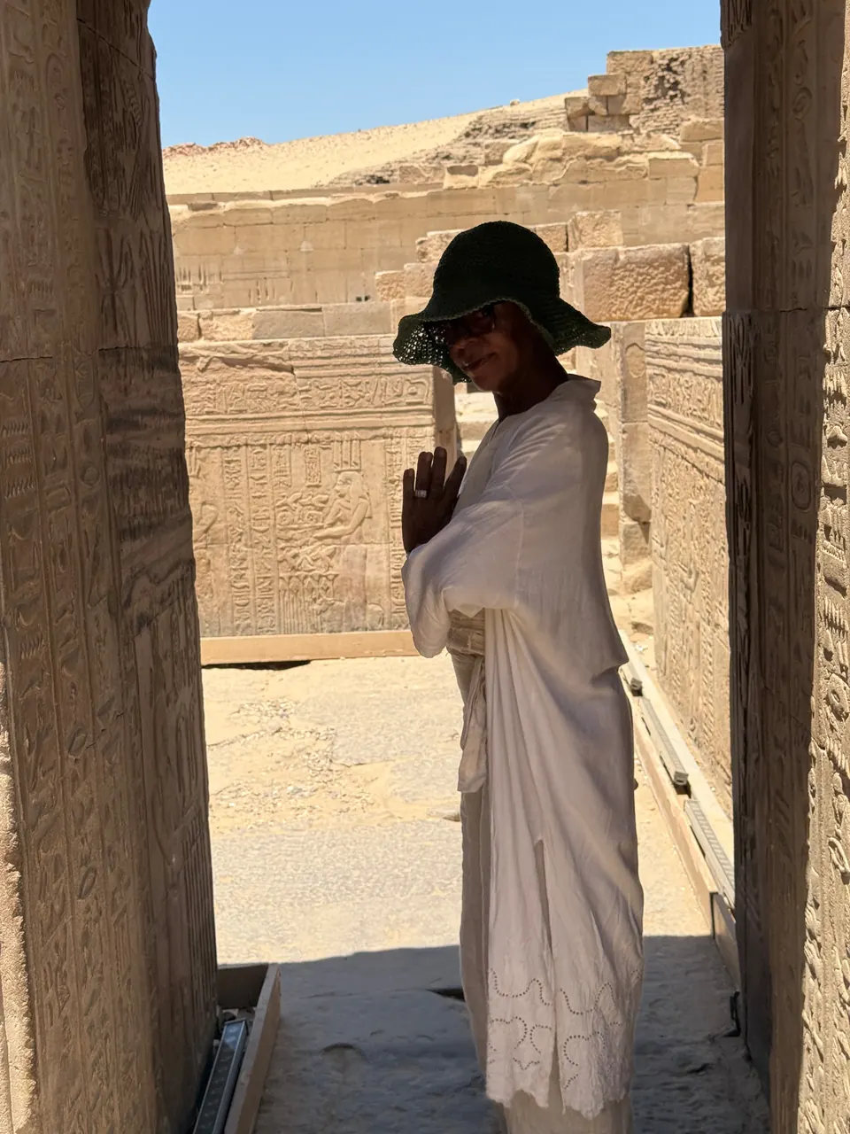 A person in white clothing and a green hat standing between carved stone columns at the Temple of Auset